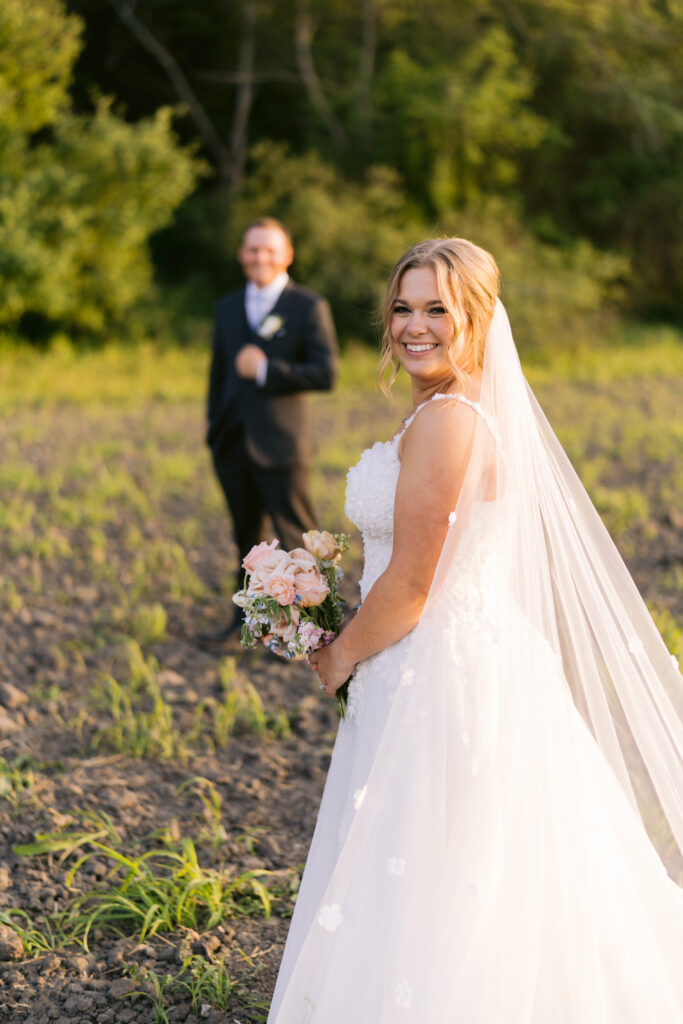 bride smiling at camera with groom in background at golden hour